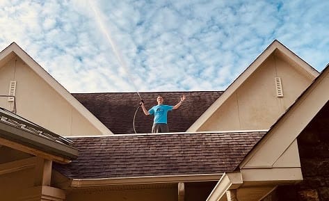 An M&J Powerwashing team member standing on a shingle roof with a pressure washer wand under a blue sky